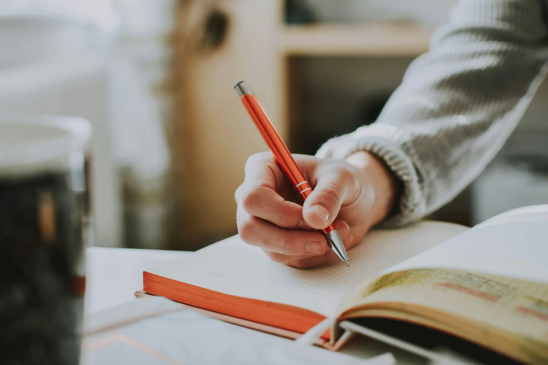 Hand writing notes with a red pen next to an open book with highlighted lines.